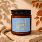 Amber glass candle with blue label reading "Loved in Every Shade VOWED".