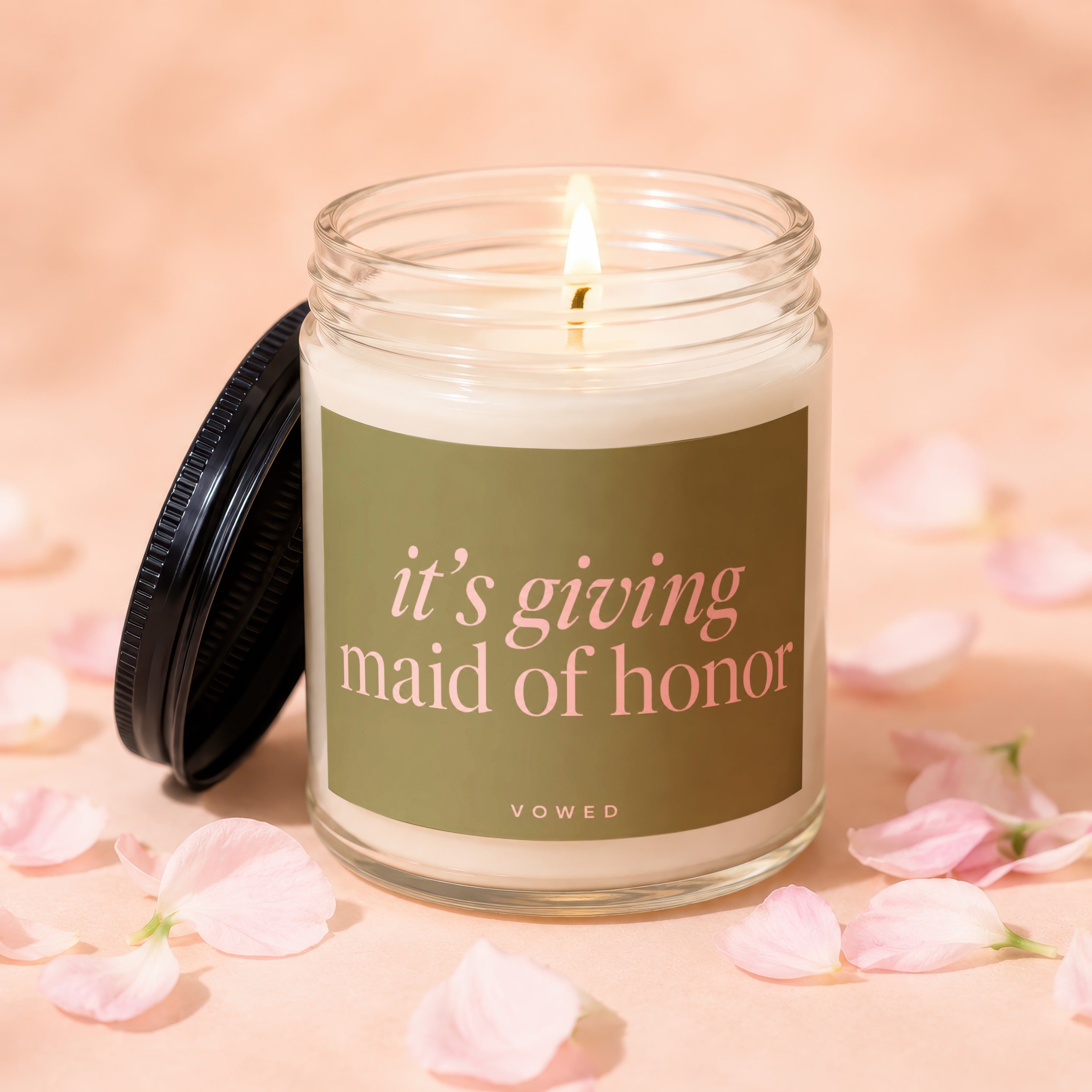 Lit jar candle on pink background with pink petals, reading "it's giving maid of honor".