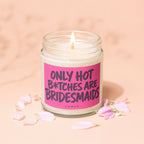 Lit candle in jar with pink label reading "ONLY HOT B*TCHES ARE BRIDESMAIDS", surrounded by petals.