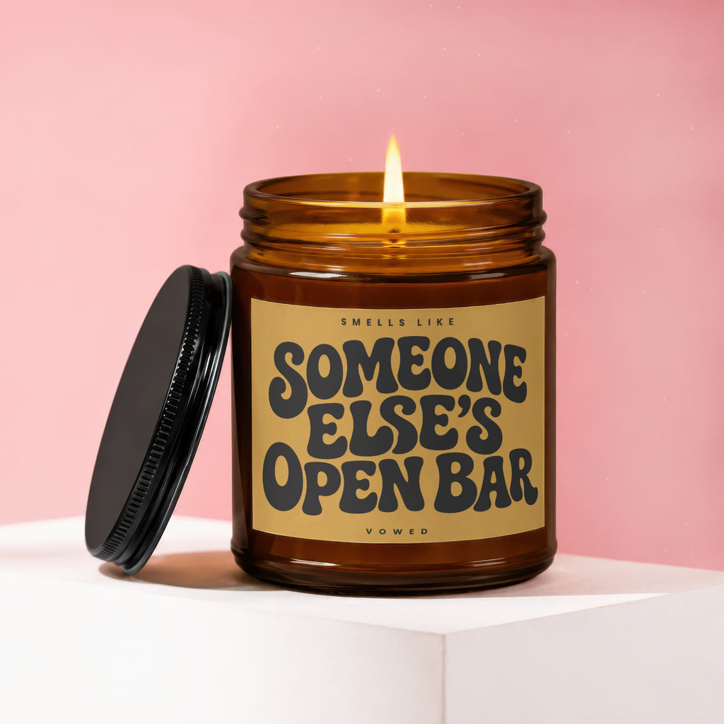 Amber jar candle with lit flame, reads "Smells like Someone Else's Open Bar."
