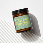 Amber glass candle with black lid and green label that reads "Our love, our rules"
