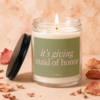 Burning candle in glass jar with "it's giving maid of honor" text on label.