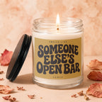 Glass jar candle with lit wick, reads "Smells Like Someone Else's Open Bar".