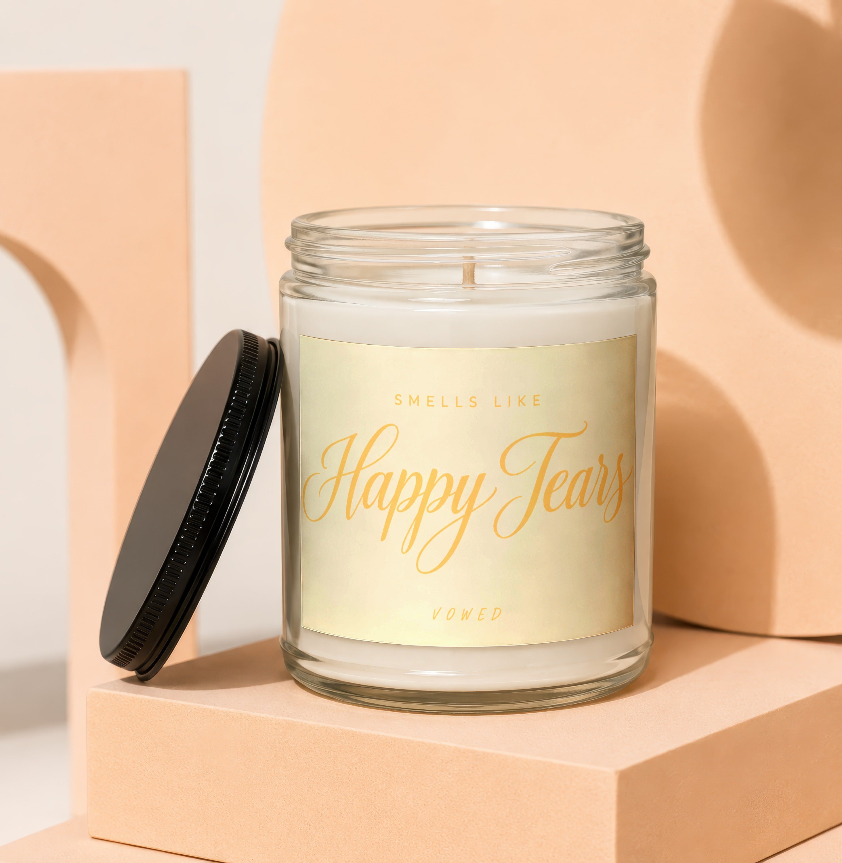 Glass candle with gold label reading "Smells Like Happy Tears Vowed" and black lid.