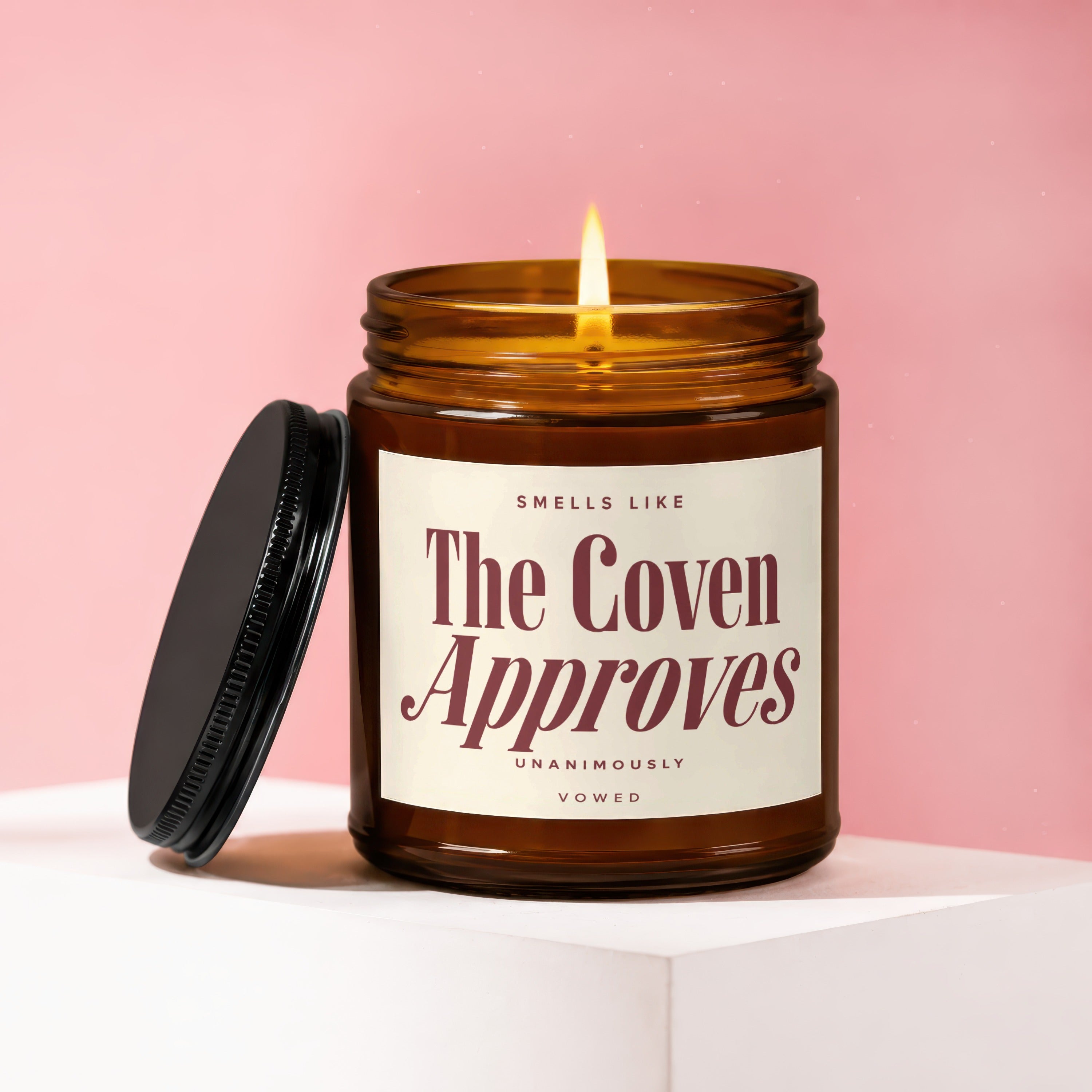 Amber glass candle with lit flame and black lid, reads The Coven Approves.