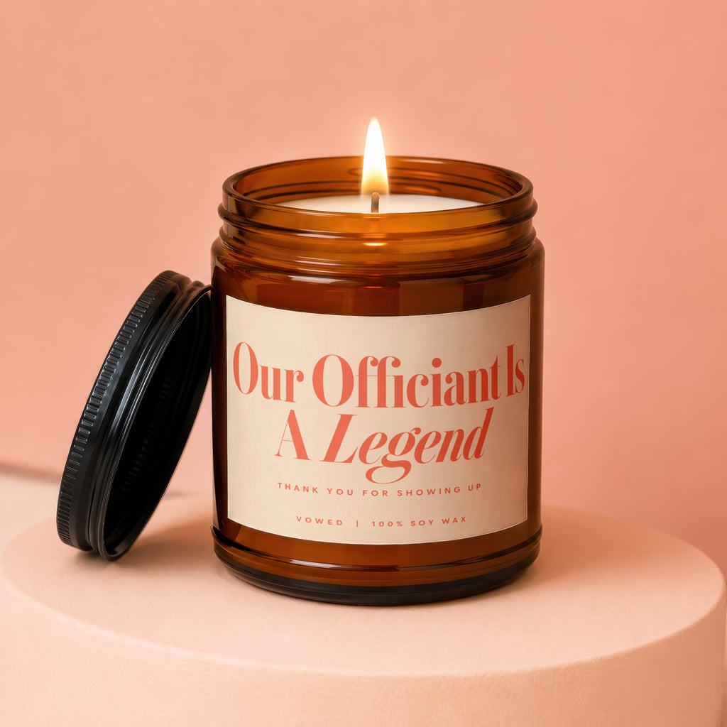 Amber jar candle with lit wick, reads "Our Officiant Is A Legend".