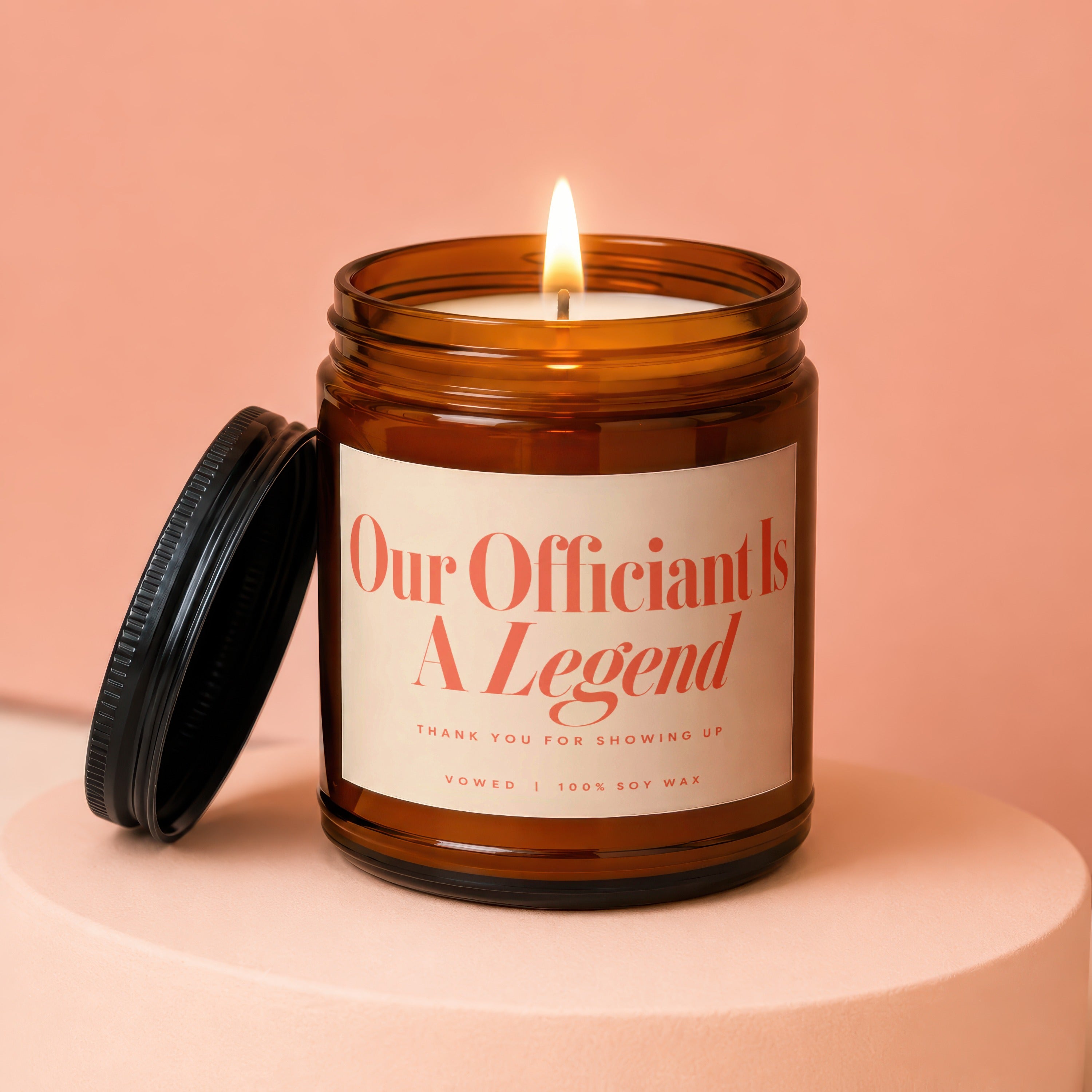 Amber jar candle with lit wick, reads "Our Officiant Is A Legend".