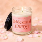 Glass candle with pink label reading "Smells Like Bridesmaid Energy" surrounded by petals.