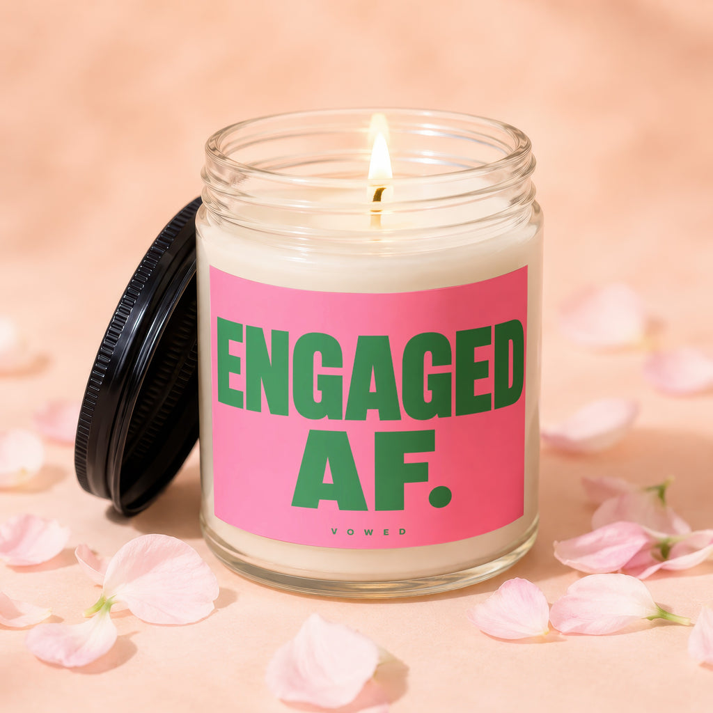 Candle with "ENGAGED AF." in green text on a pink label, lit, surrounded by petals.