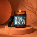 Amber glass candle with lit flame and "HE FINALLY PUT A RING ON IT" label.