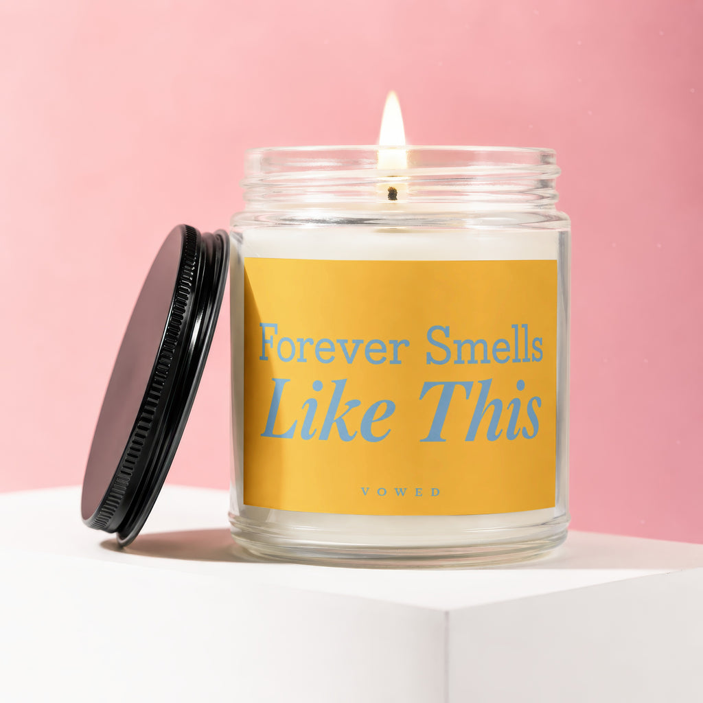 Lit candle in glass jar with yellow label reading "Forever Smells Like This Vowed"