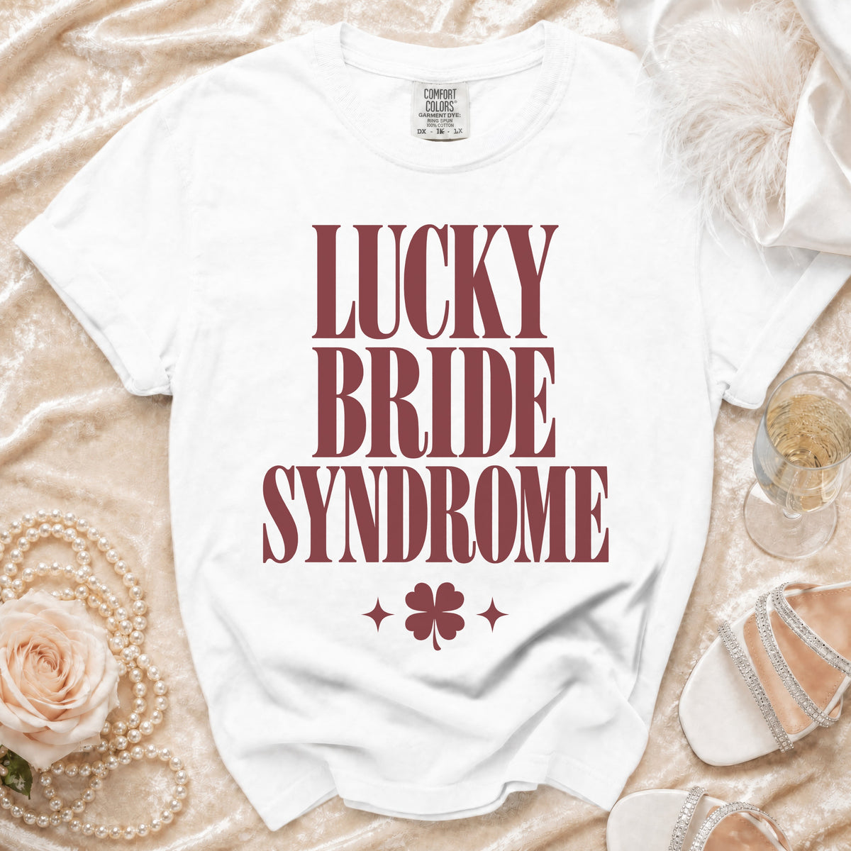 A white t-shirt with maroon text reading LUCKY BRIDE SYNDROME, surrounded by wedding-themed accessories.