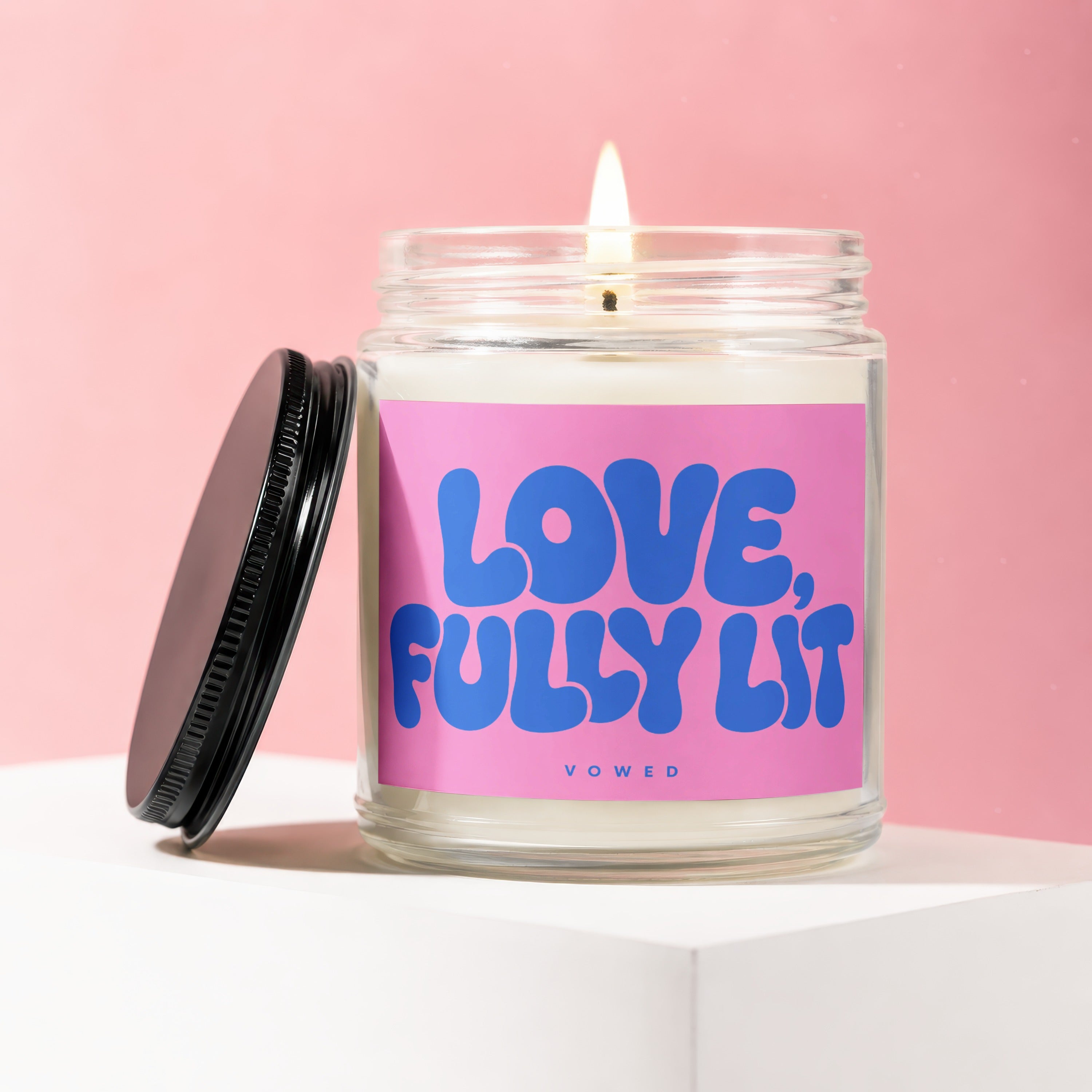 Glass candle with lit wick and pink label reading "LOVE, FULLY LIT VOWED".