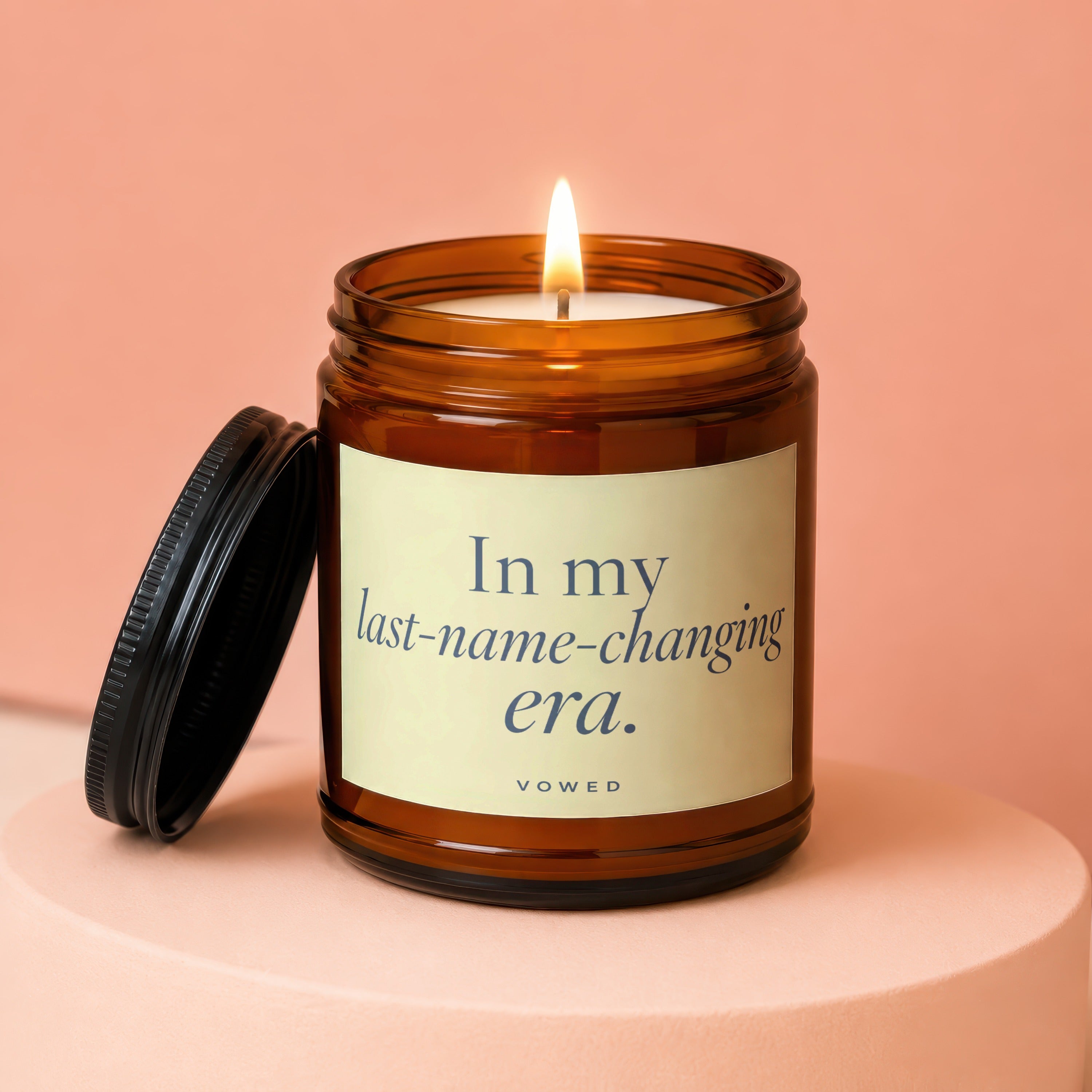 Amber jar candle with lit wick reads, "In my last-name-changing era. VOWED."