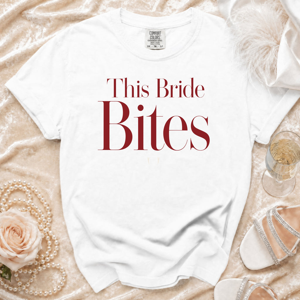 White t-shirt with "This Bride Bites" text in burgundy, styled with pearls, rose, and champagne.