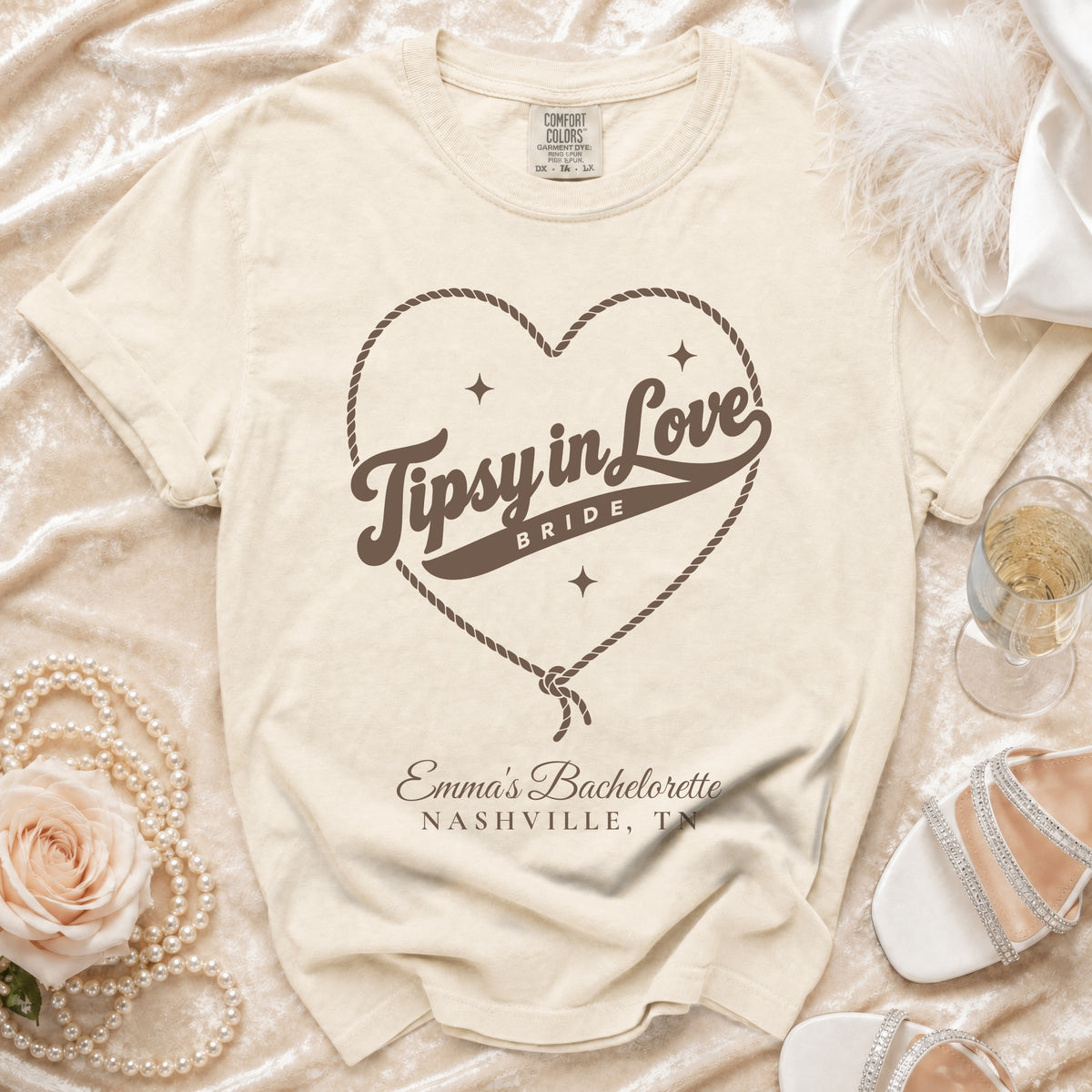 Cream t-shirt featuring a rope heart graphic, the text "Tipsy in Love Bride," and personalized bachelorette details.