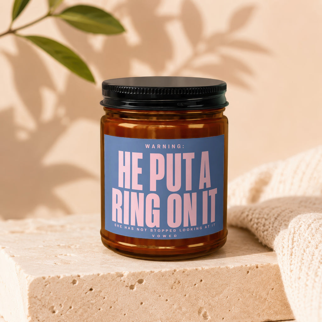 Candle with blue label reading "WARNING: HE PUT A RING ON IT (SHE HAS NOT STOPPED LOOKING AT IT) VOWED."