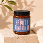 Candle with blue label reading "WARNING: HE PUT A RING ON IT (SHE HAS NOT STOPPED LOOKING AT IT) VOWED."
