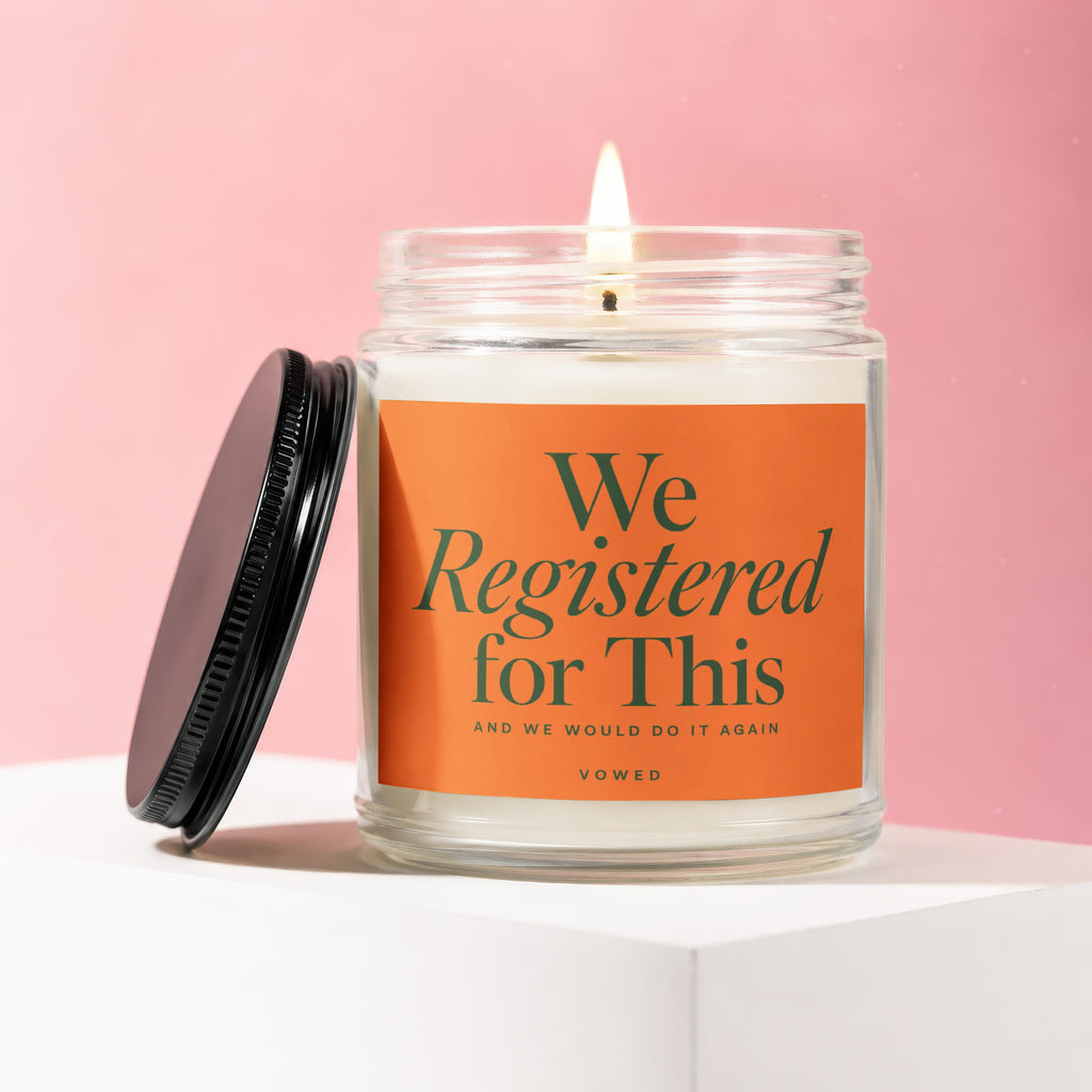 Lit candle in a glass jar with an orange label that reads "We Registered for This".