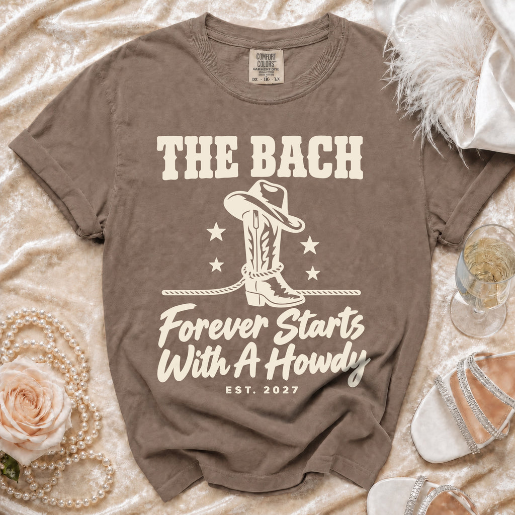 Brown short-sleeve t-shirt with cream graphics including cowboy boots, stars, and text that reads "The Bach Forever Starts With A Howdy Est. 2027".