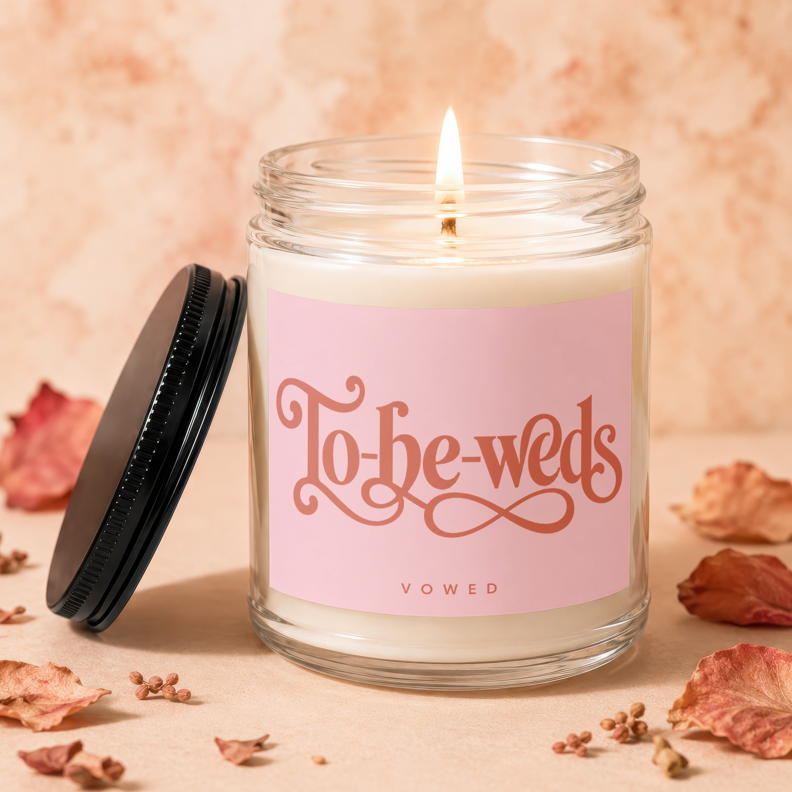 "To-be-weds" candle with pink label, lit flame, and decorative leaves.
