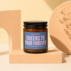 Amber glass candle with black lid, blue label reads "Cheers to your forever".