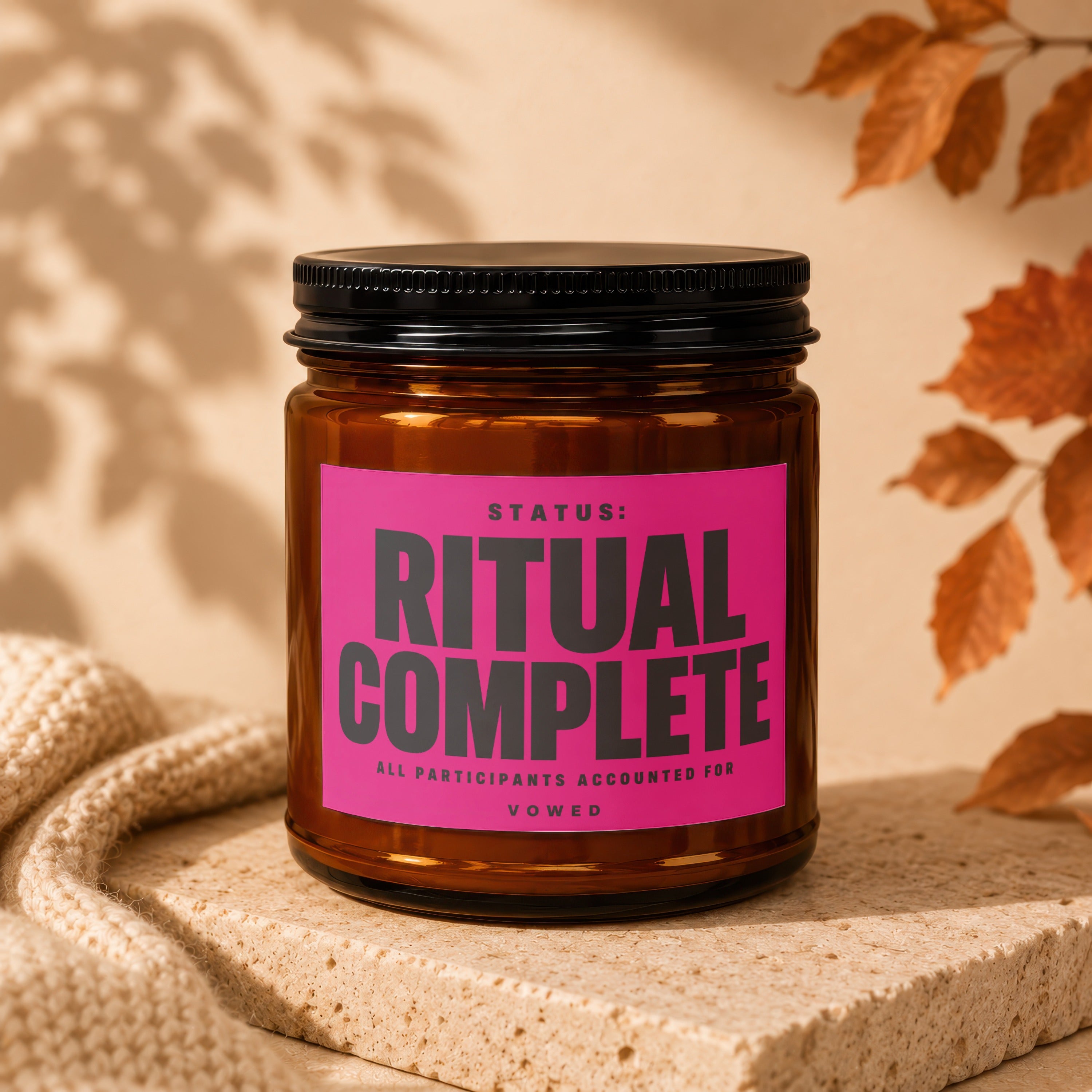Amber brown candle with pink label reading "STATUS: RITUAL COMPLETE ALL PARTICIPANTS ACCOUNTED FOR VOWED".