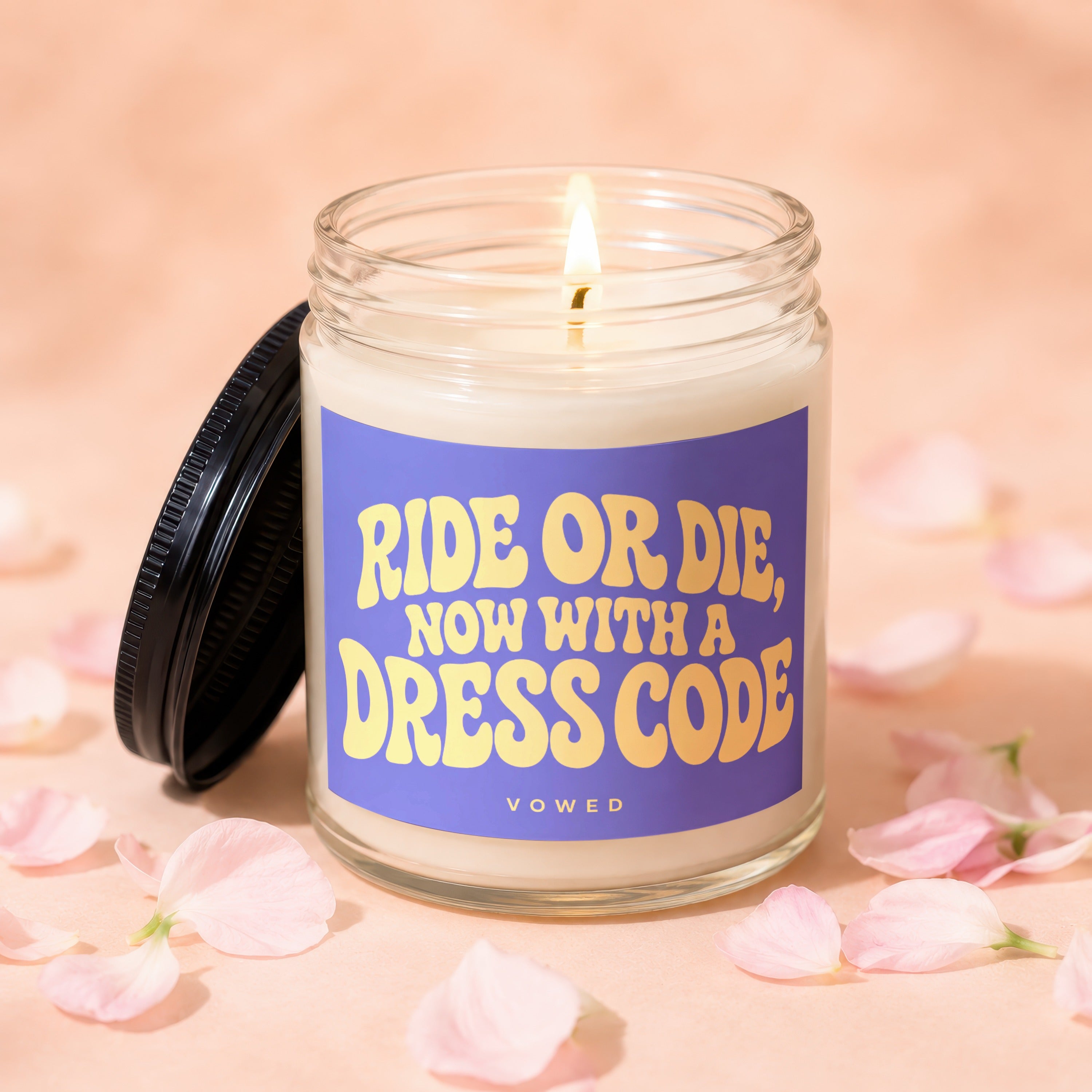 Lit candle in glass jar with purple label stating "RIDE OR DIE, NOW WITH A DRESS CODE, VOWED".