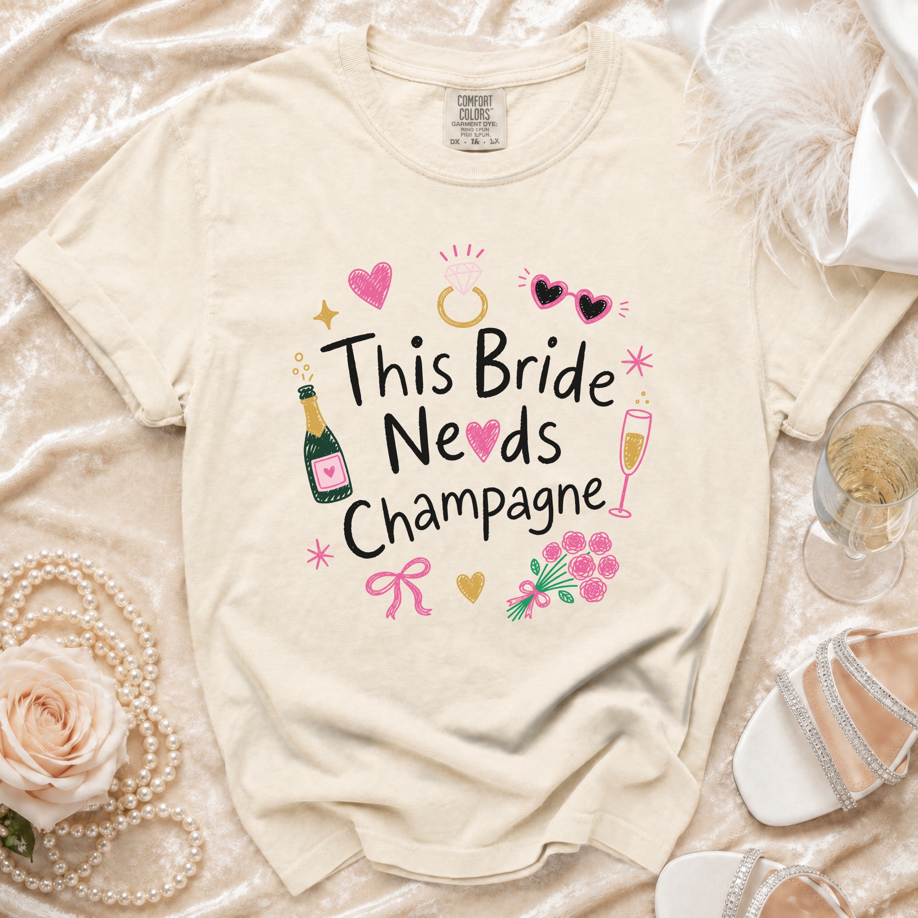 Cream t-shirt with "This Bride Needs Champagne" graphic and bridal accessories.