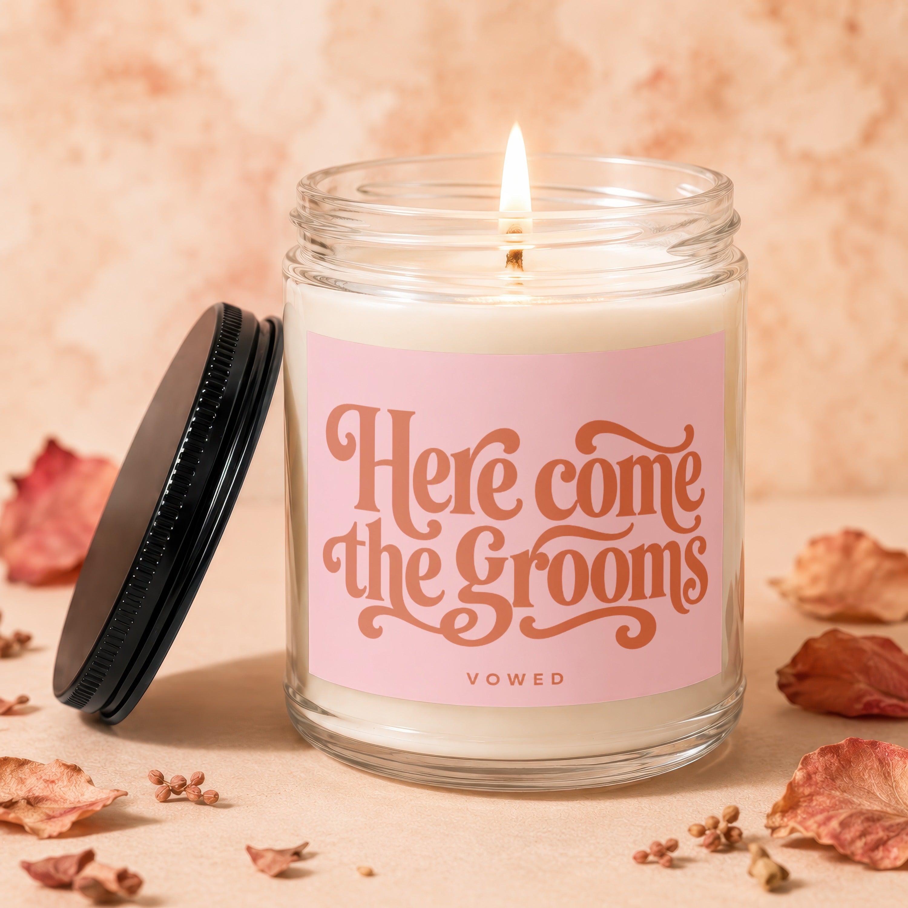 Glass candle with lit wick and pink label reading "Here come the grooms" and "VOWED".