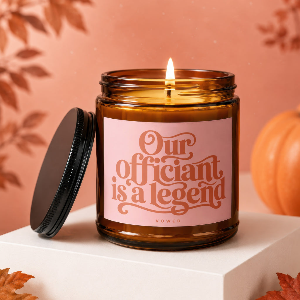 Brown glass candle with lit wick and pink label reading "Our Officiant is a Legend Vowed."
