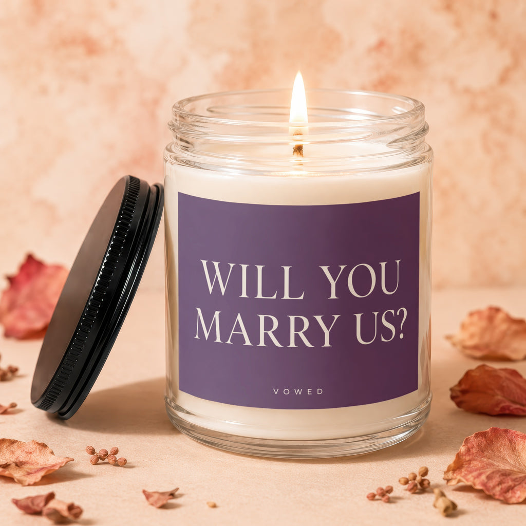 Lit candle in a glass jar with purple label reading "WILL YOU MARRY US? VOWED".