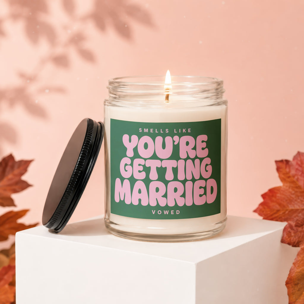 A lit candle with pink text on a green label that says "SMells LIKE YOU'RE GETTING MARRIED VOWED".