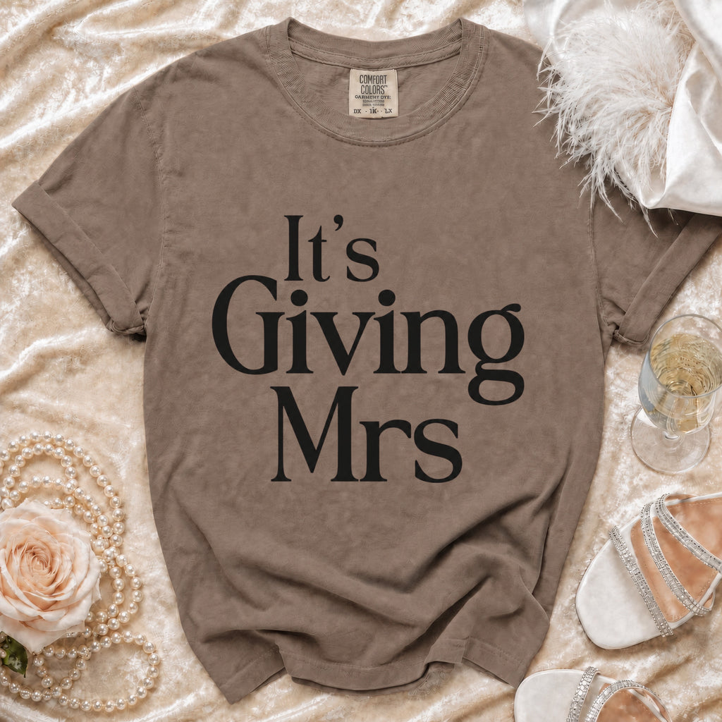 Brown t-shirt with "It's Giving Mrs" text, pearl necklace, rose, and champagne.