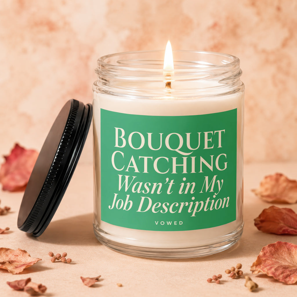 A lit candle in a glass jar with a green label reading "BOUQUET CATCHING Wasn't in My Job Description".