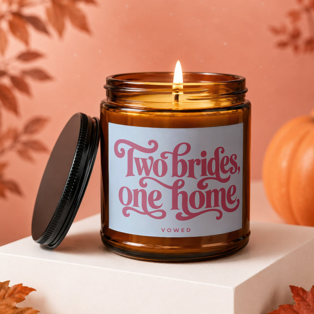 Amber jar candle with lit flame and "Two brides, one home" text on label.