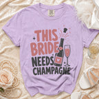 Purple t-shirt with "THIS BRIDE NEEDS CHAMPAGNE" text and champagne bottle graphic.
