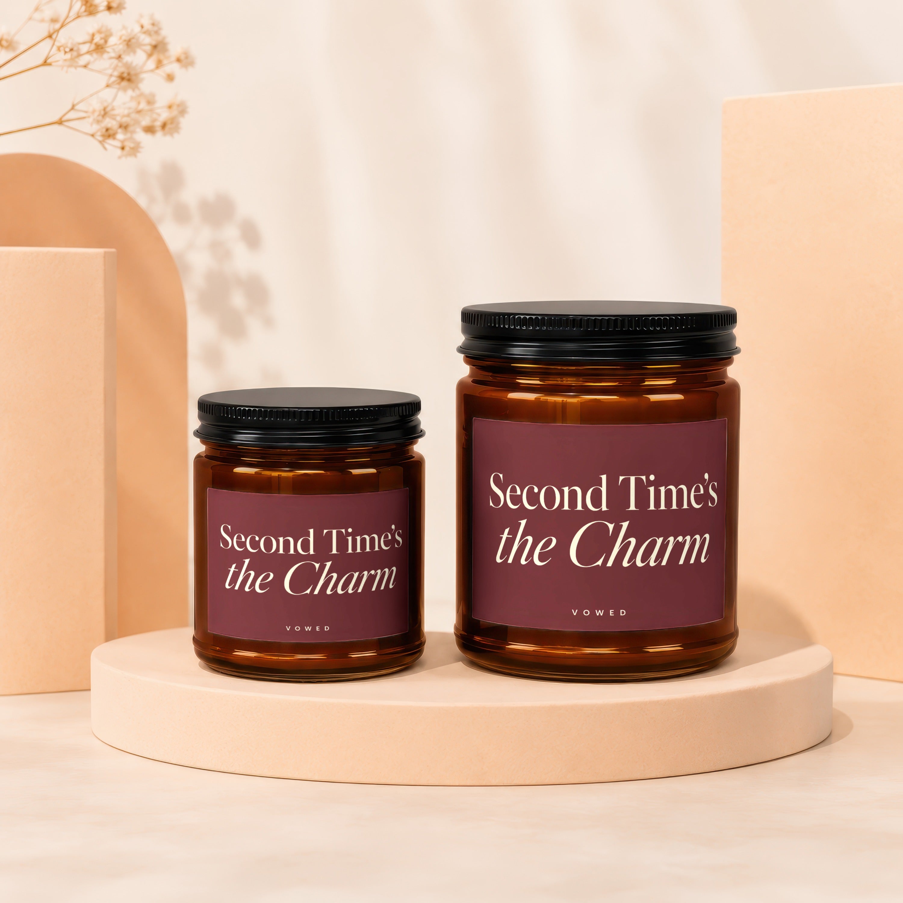 Two amber glass candles with black lids, featuring maroon labels reading "Second Time's the Charm".