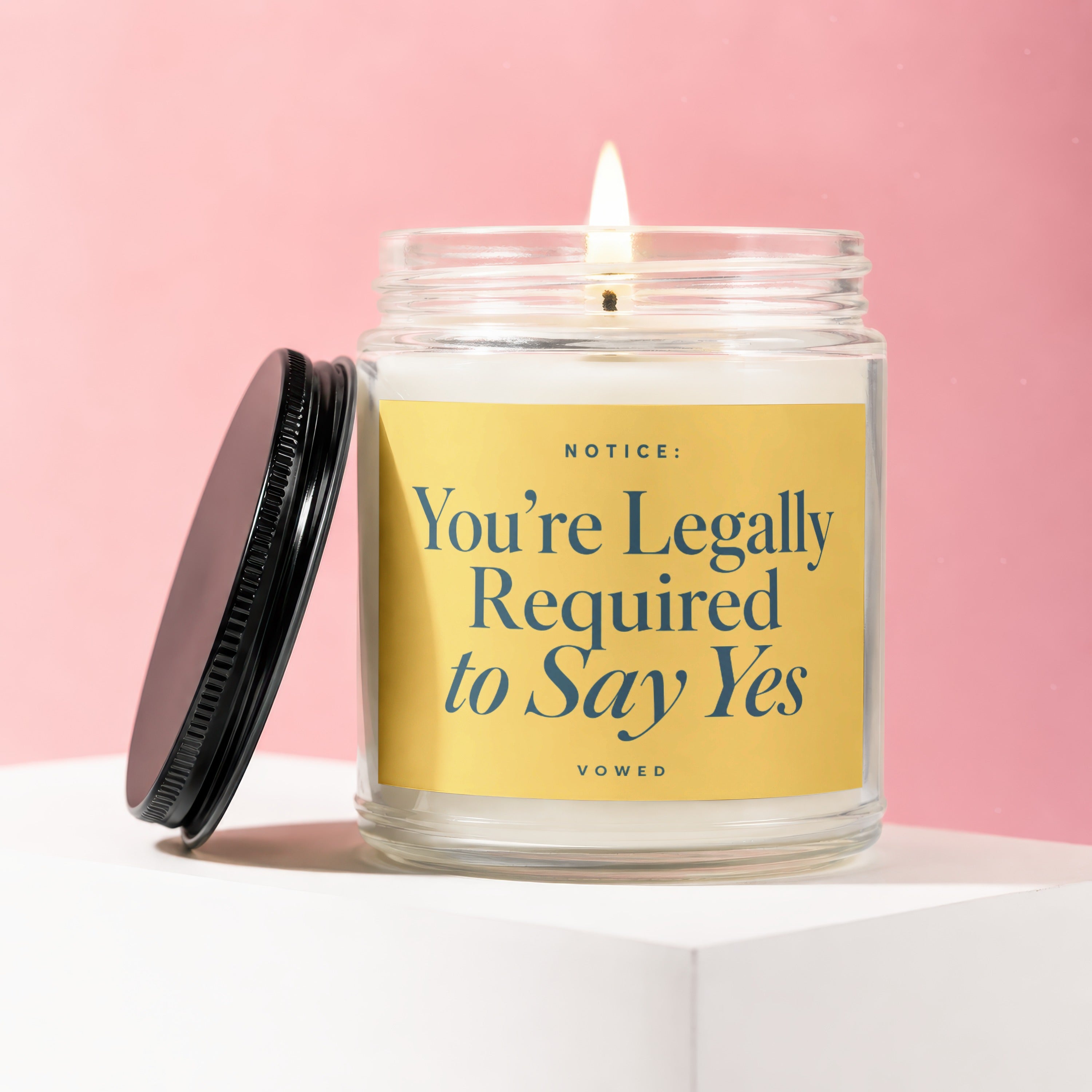 Glass candle with lit flame and yellow label reading "Notice: You're Legally Required to Say Yes".