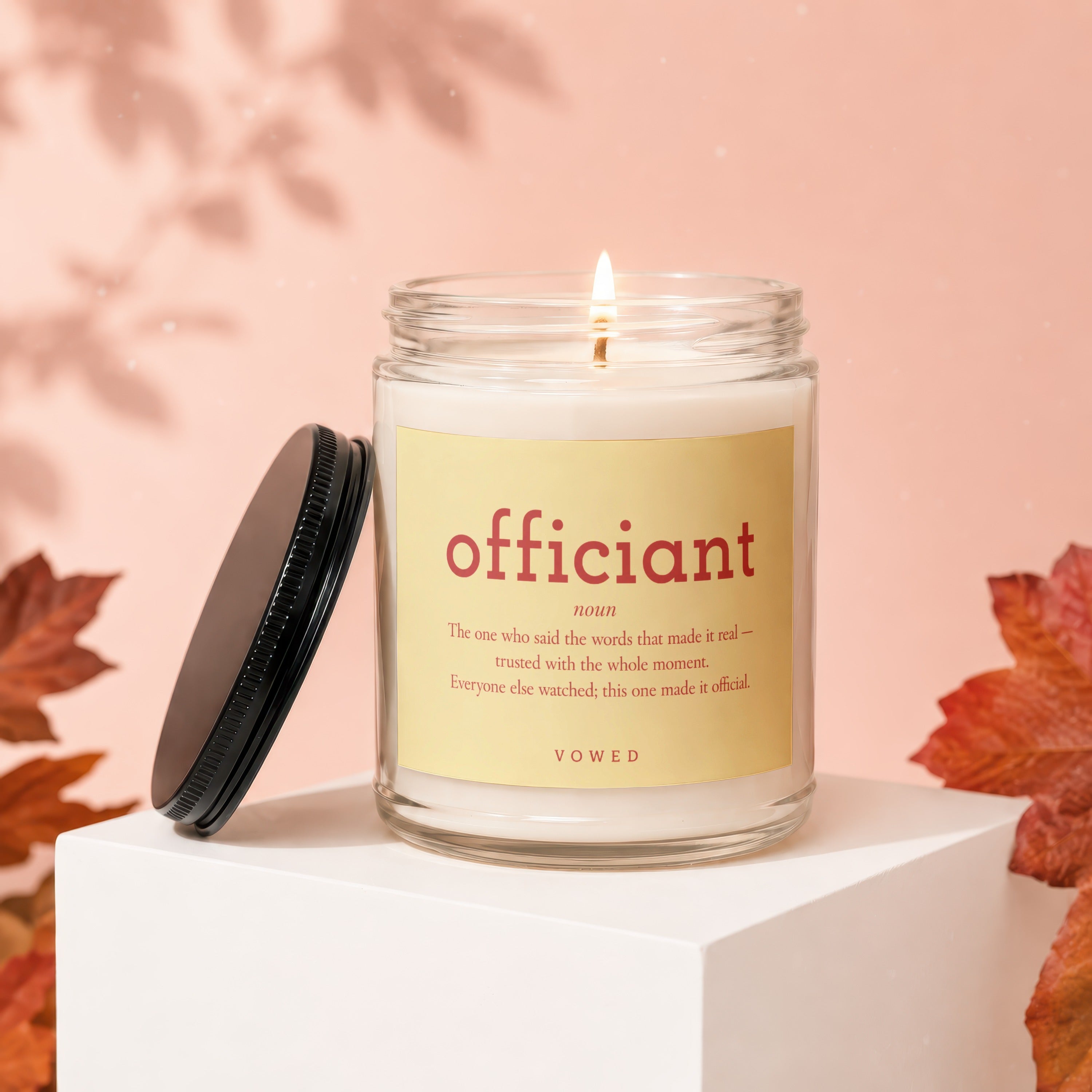 Glass jar candle with "officiant" definition label and a lit wick.