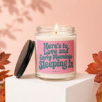 Clear glass candle with pink label reading "Here's to Love and Sleeping In".