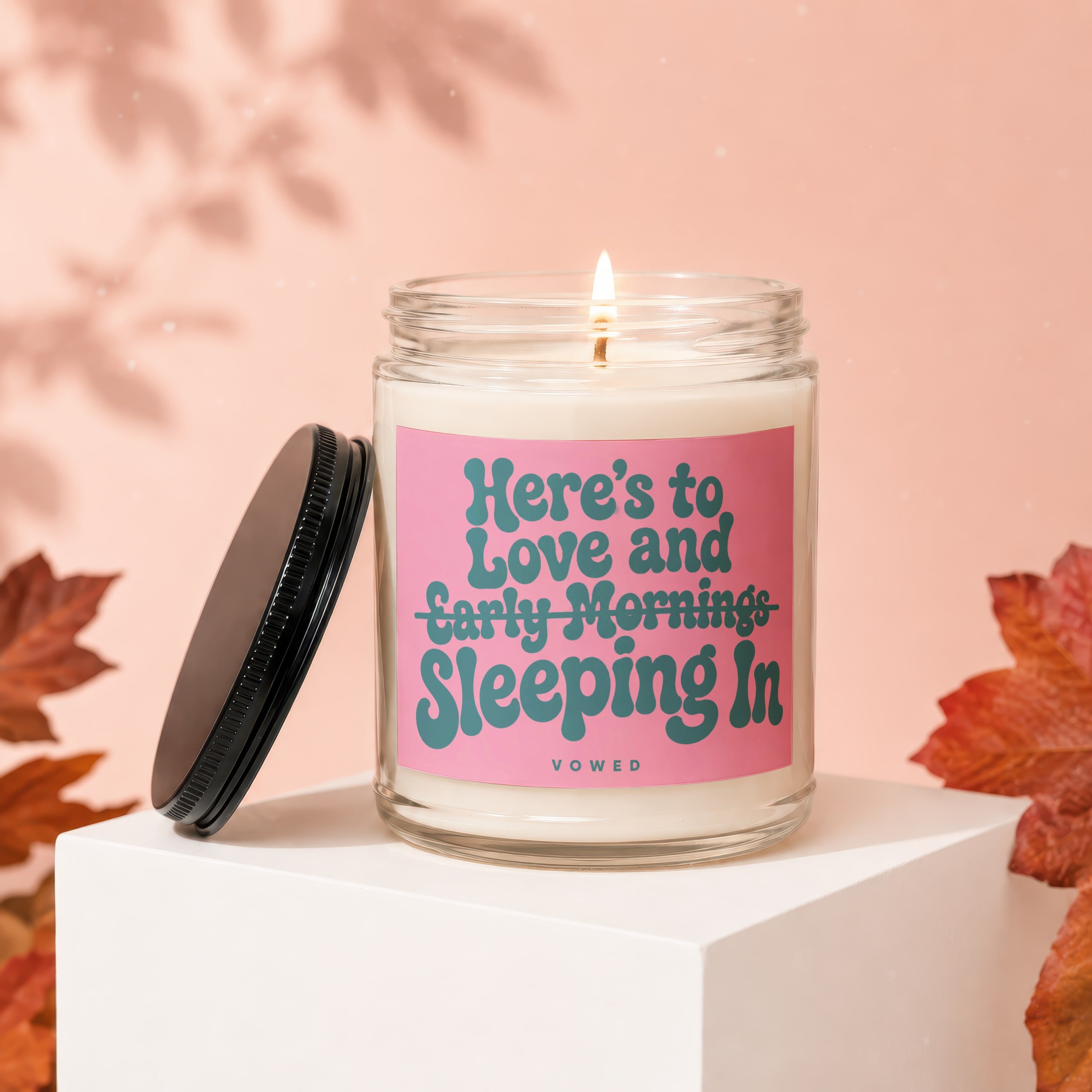 Clear glass candle with pink label reading "Here's to Love and Sleeping In".
