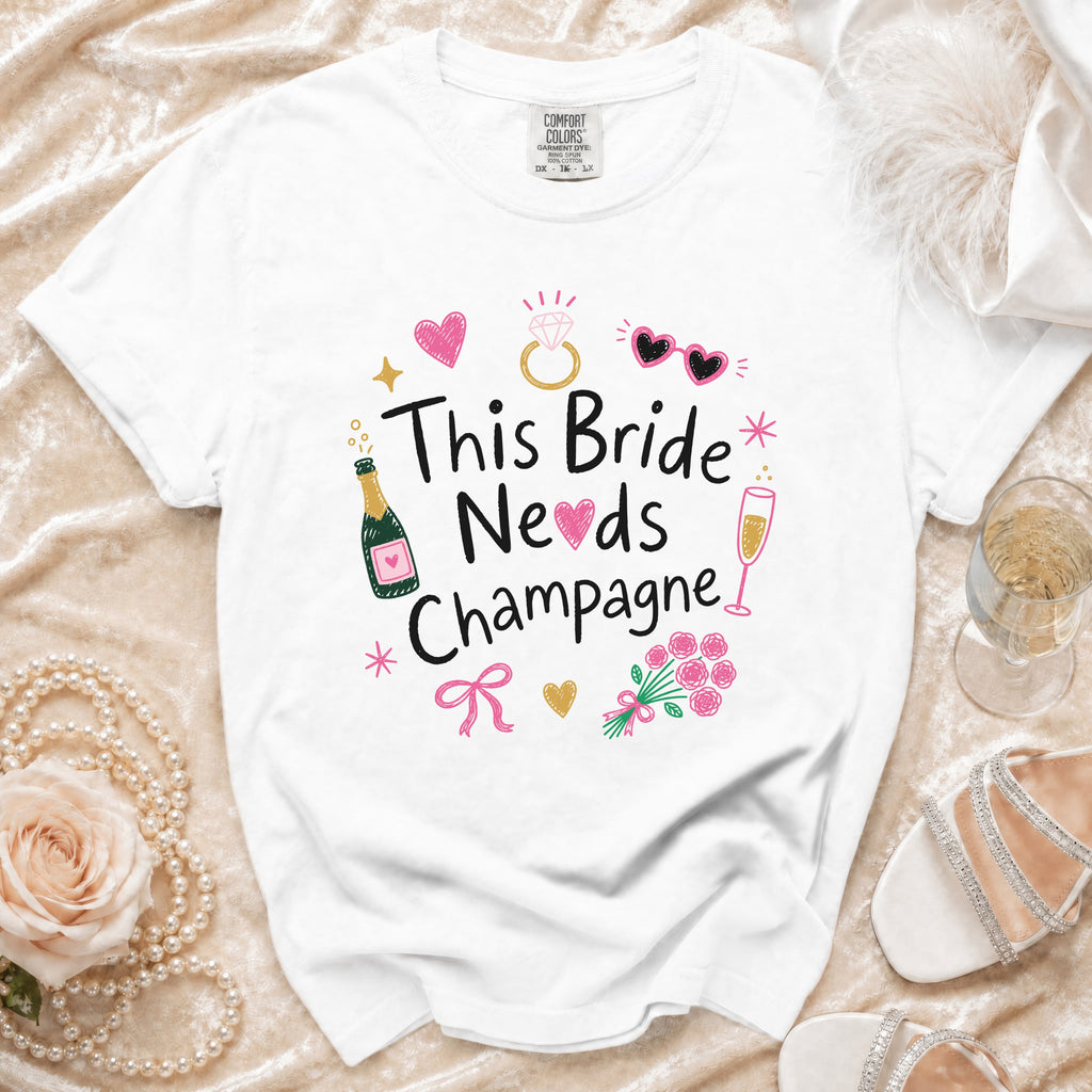 White t-shirt with "This Bride Needs Champagne" graphic, champagne bottles, and wedding icons.