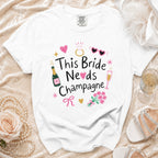 White t-shirt with "This Bride Needs Champagne" graphic, champagne bottles, and wedding icons.
