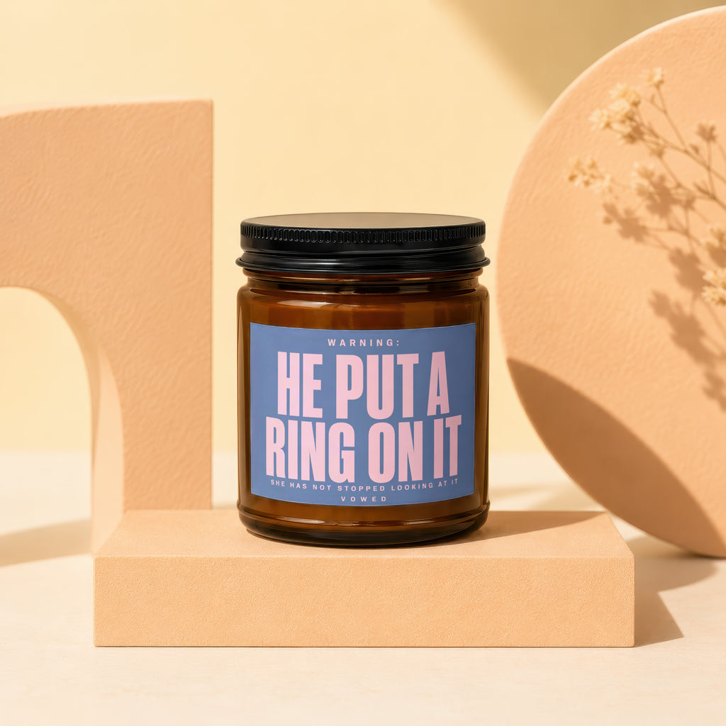 Brown glass candle with blue label text "HE PUT A RING ON IT".