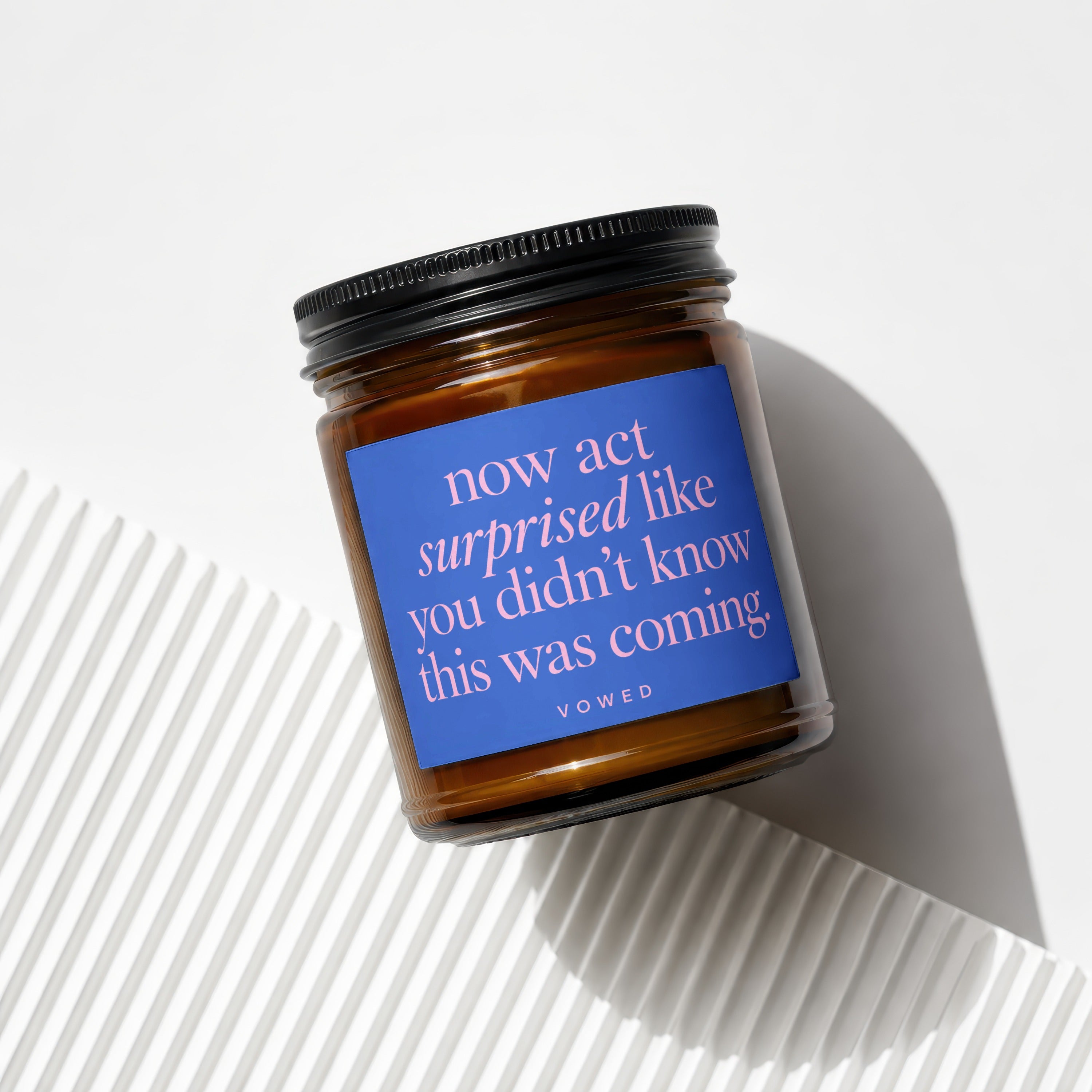 Amber jar candle with blue label reading "now act surprised like you didn't know this was coming".