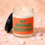 Candle with "My Hostages Bridesmaids" text on orange label, lit wick, surrounded by petals.