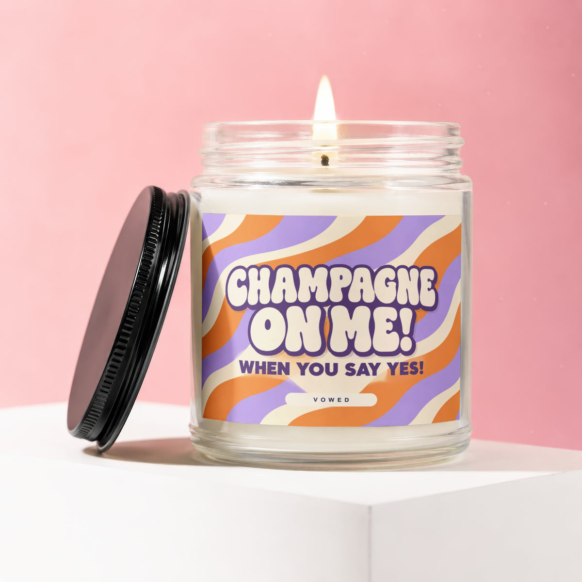 Glass candle with burning wick, featuring "Champagne On Me!" text.