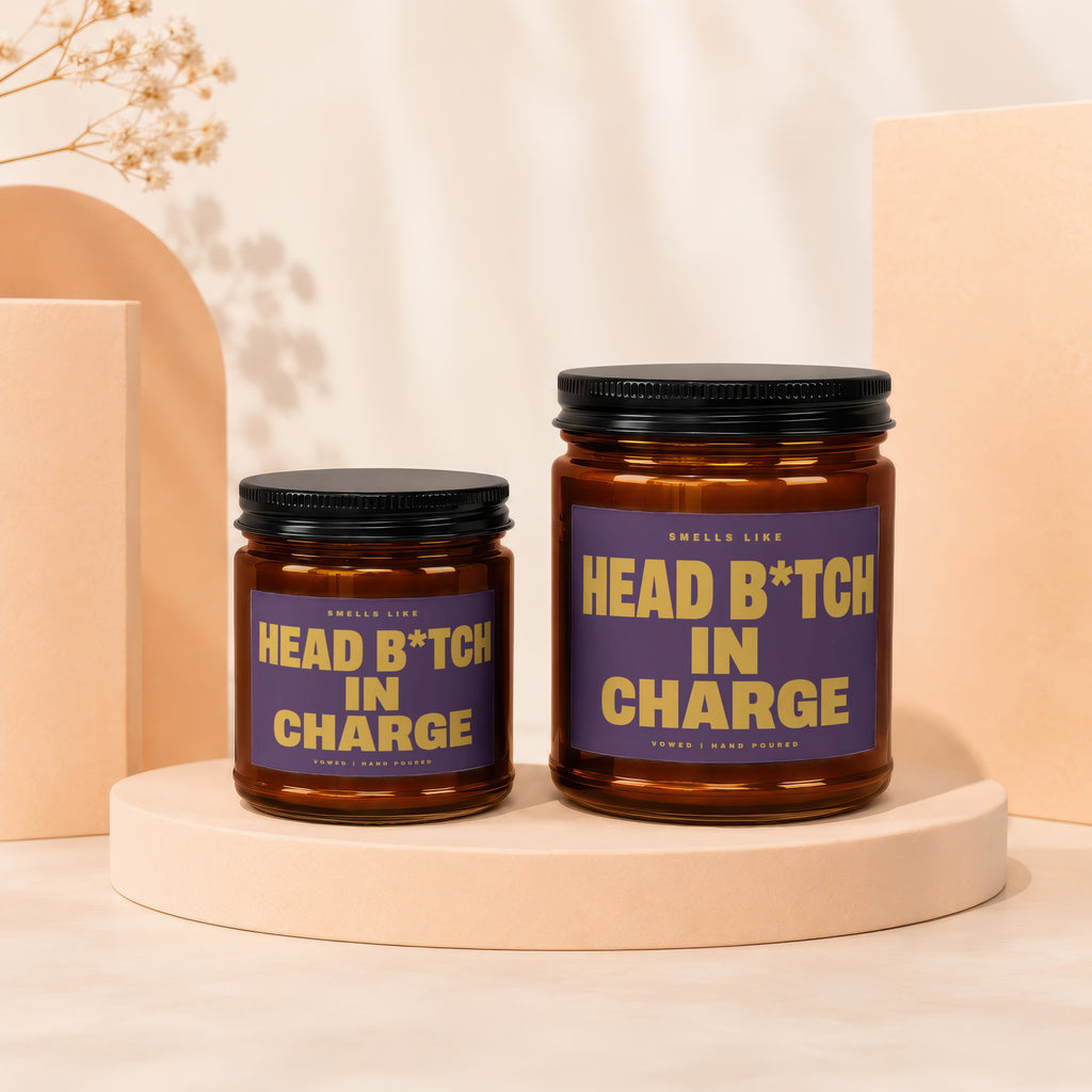 Two amber glass candles with purple labels reading "Smells Like Head B*tch In Charge".