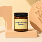 Brown glass candle with black lid and yellow label reading "Our Next Chapter, Together."
